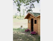 Piccola casetta in legno con tetto a due falde verde, porta aperta e finestra laterale, situata ai margini di un prato in campagna. Rami di un albero incorniciano la parte superiore e ai piedi della parete ci sono aiuole con fiori rosa e un vaso su un basamento in pietra.