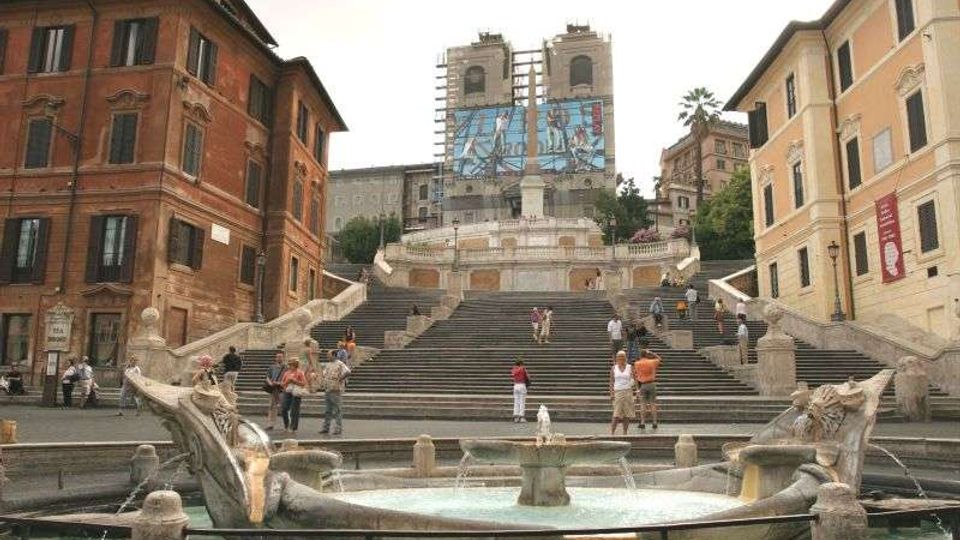 Ampia scalinata monumentale in una piazza urbana, con fontana circolare in primo piano e edifici storici ai lati. In cima alla scalinata si vede la facciata di una chiesa in restauro con impalcature e teli decorati.