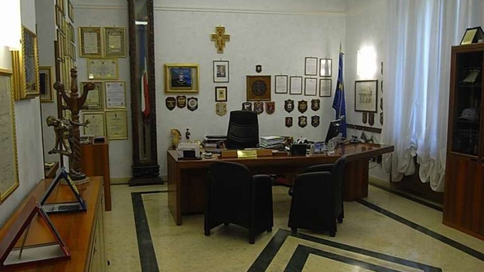 Interno di un ufficio direzionale con grande scrivania in legno, sedie nere e pareti decorate con cornici, attestati, stemmi e una bandiera vicino alla finestra con tende bianche. Pavimento in marmo chiaro con intarsi geometrici scuri e soffitto ribassato con illuminazione integrata.