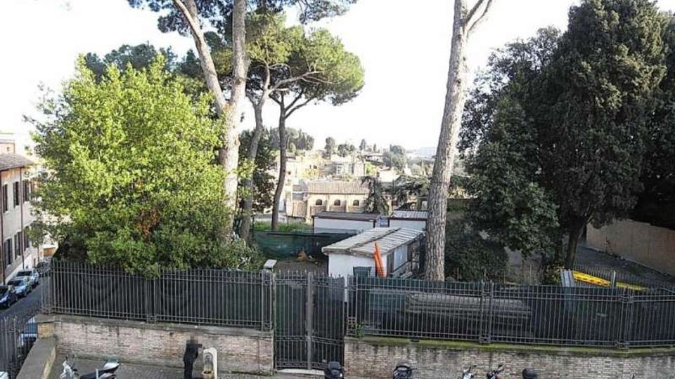 Parcheggio urbano con pavimentazione in sanpietrini e una fila di scooter lungo un muro in mattoni con recinzione metallica e cancello. Dietro la recinzione si vedono alti pini e altre alberature, con edifici e tetti della città sullo sfondo.