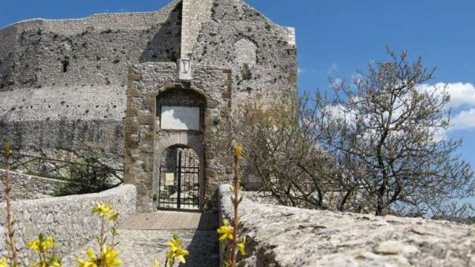 Passerella in pietra con parapetti che conduce a un ingresso ad arco con cancello metallico, inserito in mura di una fortificazione in pietra parzialmente in rovina. Intorno si vedono rocce, cespugli e piccoli alberi, con cielo azzurro sullo sfondo.