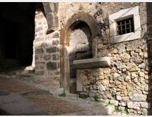 Facciata di un edificio in pietra con arco murato e portale ad arco, affiancato da una piccola finestra con inferriate. In primo piano si vede una strada lastricata in lieve salita e, a sinistra, un passaggio ad arco che conduce in una zona in ombra.