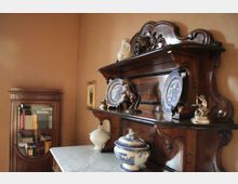 Sala d’interni con mobili in legno scuro in stile classico, tra cui una credenza con ripiani decorati con piatti in ceramica e statuette e un mobile vetrina nell’angolo. In primo piano si vede un tavolo con piano in marmo e un vaso in ceramica blu e bianca.