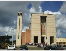 Facciata di una chiesa moderna in pietra chiara con grandi portali d’ingresso e un alto mosaico verticale al centro, affiancata da un campanile slanciato con croce in cima. Davanti si apre una piazza con auto parcheggiate e lampioni, sotto un cielo nuvoloso.