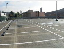 Parcheggio all’aperto su un piano rialzato, con ampie strisce bianche di delimitazione dei posti e cordoli di gomma. Sullo sfondo si vedono una ringhiera, edifici bassi, alberi e alcuni lampioni sotto un cielo chiaro.