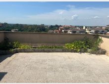 Terrazza panoramica con pavimento piastrellato e muretto perimetrale, con una lunga fioriera in cemento con vegetazione. Sullo sfondo si vede un quartiere di case e alberi, con colline lontane sotto un cielo azzurro con nuvole.