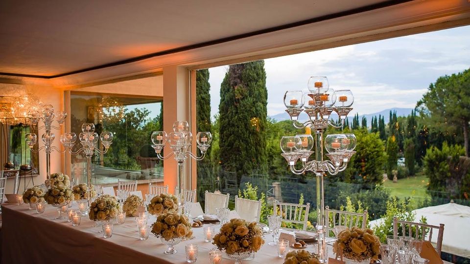 Sala ricevimenti con ampia vetrata panoramica affacciata su un giardino con cipressi e colline sullo sfondo. Lungo tavolo apparecchiato con tovaglia bianca, composizioni floreali, bicchieri e candelabri in vetro trasparente.