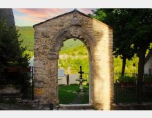 Antico portale in pietra con arco scolpito e decorazioni, che introduce a un piccolo giardino. Oltre l’arco si vede una fontana centrale e, sullo sfondo, colline verdi con alberi e un cielo al tramonto.