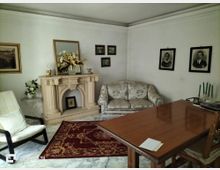 Soggiorno con pareti bianche e pavimento in marmo, con camino decorativo in pietra e quadri appesi. Nella stanza ci sono un divano a due posti, una poltrona, un grande tavolo in legno e un tappeto rosso con motivi floreali.