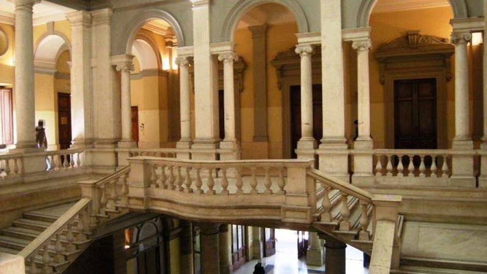 Interno di un edificio storico con cortile a doppia altezza, caratterizzato da colonne e arcate in pietra. Una scala monumentale con balaustre conduce a un ballatoio superiore affacciato sull’atrio con pavimento lucido e grandi porte.