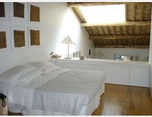 Camera da letto mansardata con pavimento in legno, letto matrimoniale con biancheria chiara e pareti bianche decorate con quadri. Il soffitto è in travi di legno a vista con lucernario, e lungo il lato c’è un mobile basso con lampada.