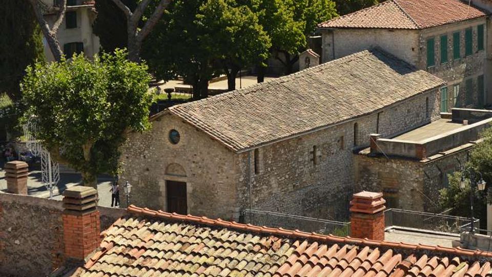 Vista dall’alto di un complesso di edifici in pietra con tetti in coppi e tegole, tra cui una struttura rettangolare con facciata semplice e piccola finestra circolare. Intorno si vedono alberi, una piazzetta lastricata e muretti con ringhiere ai margini.