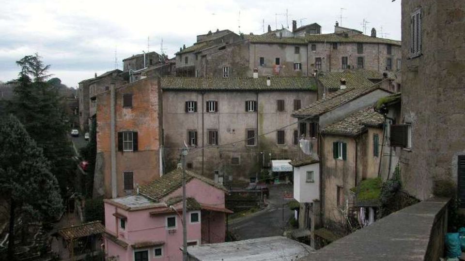 Veduta di un borgo collinare con edifici in muratura addossati tra loro e tetti in tegole, affacciati su una strada stretta. In primo piano si vedono un muretto e piccole case, mentre sullo sfondo compaiono alberi e un cielo nuvoloso.