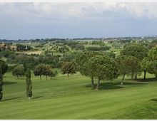 Ampio campo da golf con prati verdi ondulati e alberi sparsi, con alcuni cipressi in primo piano. Sullo sfondo si vede una campagna pianeggiante con edifici lontani sotto un cielo parzialmente nuvoloso.