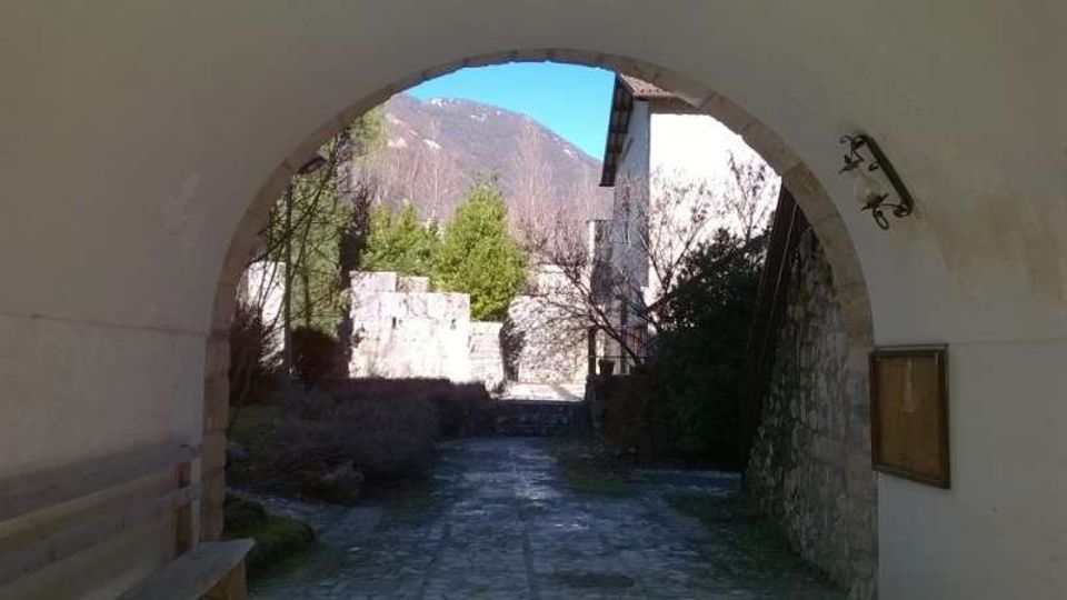Passaggio ad arco in muratura che incornicia un vialetto acciottolato verso un cortile con edifici in pietra e vegetazione. Sullo sfondo si vedono alberi e montagne, con una parete in pietra e una lampada a muro lungo il lato destro.