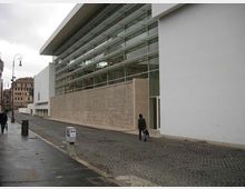 Edificio moderno con lunga facciata vetrata a più livelli e ampie pareti bianche, affacciato su una strada in pavé. L’ingresso è vetrato e sullo sfondo si vedono lampioni e palazzi urbani.