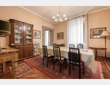 Sala da pranzo classica con grande tavolo rettangolare coperto da tovaglia e sei sedie, su tappeto persiano e pavimento in parquet. Lungo le pareti ci sono una credenza in legno con vetrina, quadri e una lampada da terra, con lampadario a soffitto e alta finestra con tende chiare.