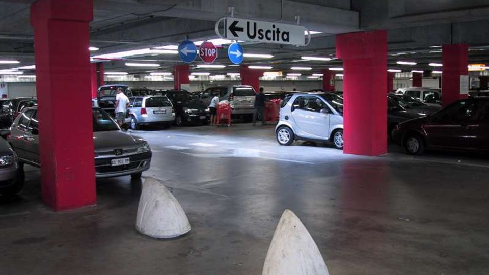 Parcheggio sotterraneo multipiano con colonne strutturali rosse, file di auto in sosta e illuminazione a neon sul soffitto in cemento. Al centro sono visibili cartelli stradali sospesi con la scritta “Uscita” e indicazioni di direzione.