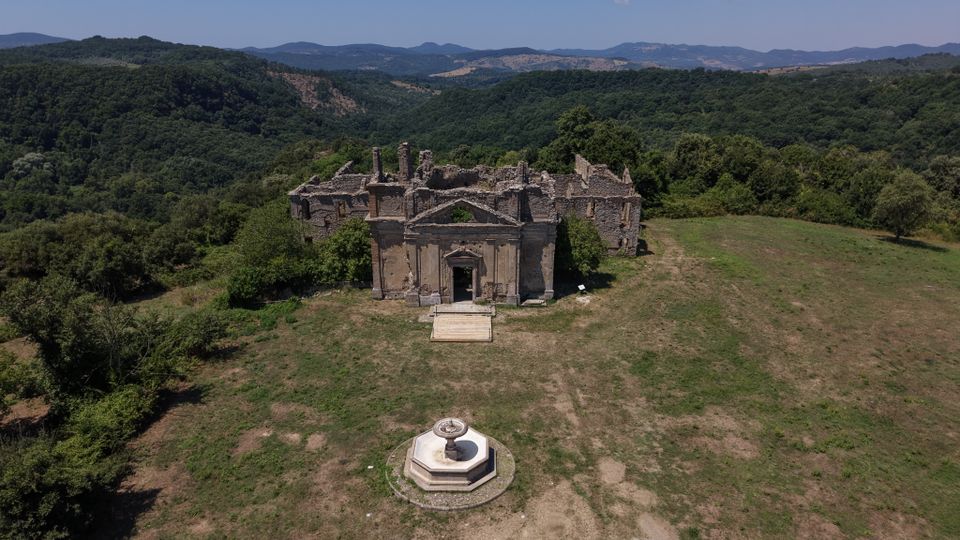 Veduta aerea di un edificio storico in rovina con facciata in pietra e ingresso centrale, isolato in una radura. In primo piano si vede una fontana ottagonale, con boschi e colline montuose sullo sfondo sotto un cielo sereno.