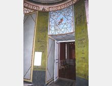 Interno di un edificio storico con grande portale decorato, pareti verdi con pannelli geometrici e fregi dipinti. Sopra l’ingresso un affresco con un uccello entro motivi ornamentali, che conduce a una sala adiacente con pavimento scuro e sedie lungo la parete.