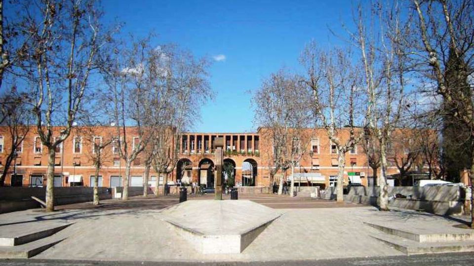 Piazza urbana pavimentata con gradoni e una piattaforma centrale rialzata, circondata da alberi spogli. Sullo sfondo si vede un grande edificio in mattoni rossi con una facciata simmetrica e un porticato ad archi.