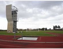 Campo sportivo all’aperto con pista di atletica rossa e prato centrale con porte da calcio. A sinistra si vede una torre alta per arrampicata o allenamento con struttura metallica, sotto un cielo nuvoloso e con cavi sospesi sopra l’area.