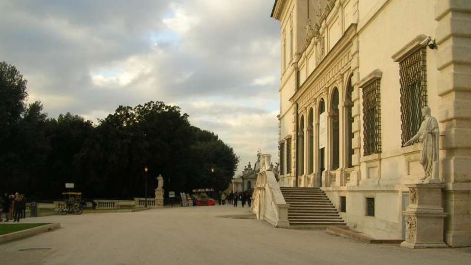 Viale pedonale accanto a un grande edificio storico in pietra chiara, con facciata monumentale, finestre con grate e una scalinata che conduce a portici ad arco. Ai lati si vedono statue su piedistalli e, sullo sfondo, un’area verde con alberi sotto un cielo nuvoloso.