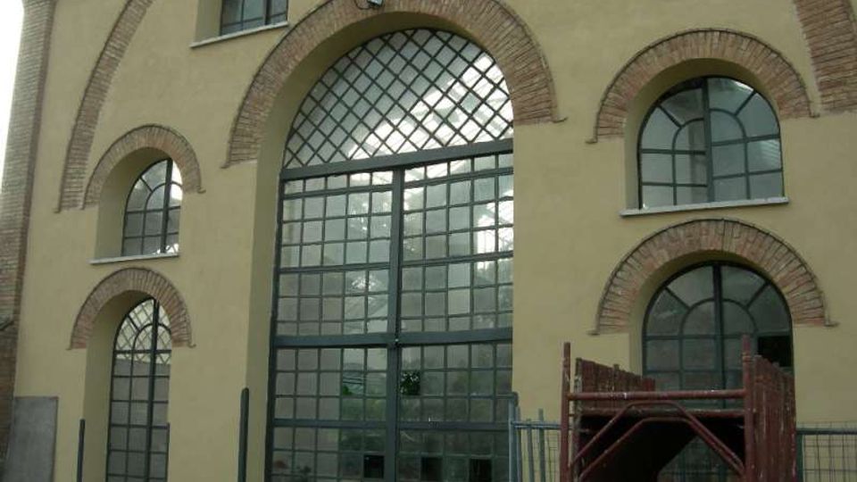 Facciata di un edificio in intonaco chiaro con grandi finestre ad arco e porta centrale vetrata a griglia, incorniciate da archi in mattoni. In primo piano si vedono una recinzione metallica e materiali da cantiere accatastati.