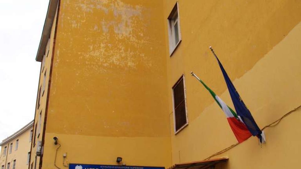 Ingresso di un edificio scolastico a più piani con facciata gialla, porta vetrata e insegna blu “Liceo Statale Ilaria Alpi”. Accanto all’entrata sono visibili una rampa con corrimano verde e due bandiere (Italia e Unione Europea) fissate al muro.