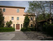 Edificio a due piani intonacato color pesca con tetto in tegole e porta d’ingresso centrale, affacciato su un cortile asfaltato. Davanti ci sono siepi e alberi, con un passaggio coperto sul lato destro accanto a una recinzione.
