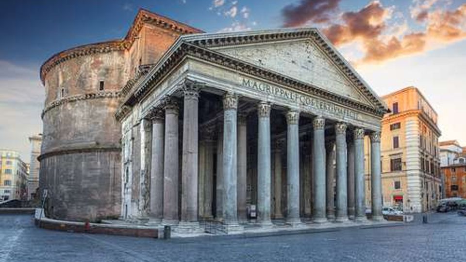 Tempio romano in pietra con grande portico a colonne e frontone triangolare, affiancato da una rotonda muraria sul retro. L’edificio si trova in una piazza lastricata, circondato da palazzi cittadini sotto un cielo al tramonto.