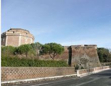 Veduta esterna di una fortificazione storica in mattoni e pietra, con bastioni inclinati e un edificio ottagonale sullo sfondo. Davanti corre una strada asfaltata con muretto e siepi, affiancata da pini sotto un cielo sereno.