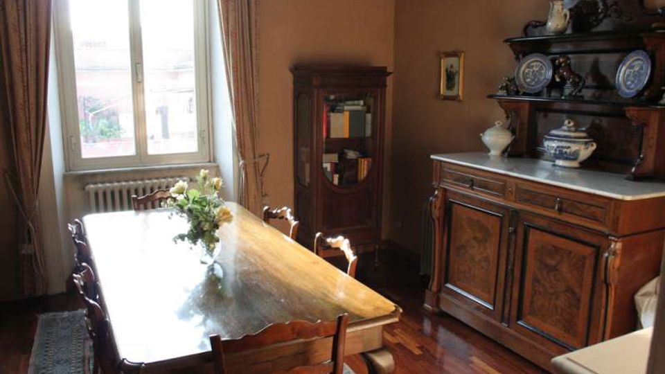 Sala da pranzo con grande tavolo in legno e sedie, illuminata da una finestra alta con tende e radiatore sottostante. Sulla destra c’è una credenza antica con alzata e piatti decorativi, e sul fondo una vetrina in legno con libri.