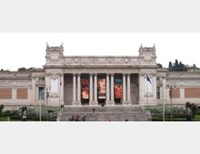 Facciata monumentale in stile neoclassico della Galleria Nazionale d’Arte Moderna, con ampio portico a colonne, grandi scalinate centrali e banner verticali appesi tra le colonne. Davanti si vede un’area verde con vialetti e lampioni, con bandiere ai lati dell’ingresso.
