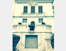 Facciata di un edificio storico a più piani con finestre simmetriche e balconata curvilinea; al centro si vede una nicchia con statua sopra un grande pannello decorativo geometrico, con vegetazione ai lati.