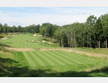 Campo da golf con fairway ampio e ondulato, delimitato da erba alta e circondato da un fitto bosco di alberi. Lungo il percorso si vedono diverse buche di sabbia bianche e leggere pendenze del terreno sotto un cielo parzialmente nuvoloso.