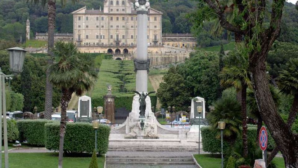 Vista di un viale monumentale in un giardino urbano con palme e siepi potate, che conduce a una grande colonna con sculture e scalinate. Sullo sfondo si vede un ampio edificio storico su una collina boscosa.
