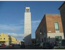 Strada urbana con edifici in mattoni e intonaco e una torre alta e rettangolare in cemento con bandiera in cima. Auto parcheggiate e in transito ai lati della via, con cielo azzurro sullo sfondo.
