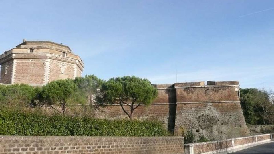 Veduta esterna di una fortificazione storica in mattoni e pietra, con bastioni inclinati e un edificio ottagonale sullo sfondo. Davanti corre una strada asfaltata con muretto e siepi, affiancata da pini sotto un cielo sereno.