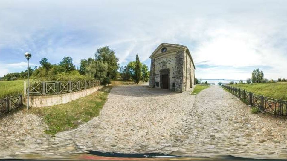 Piazzale in ciottoli accanto a una piccola chiesa in pietra con facciata a timpano e ingresso centrale. Tutt’intorno si vedono campi verdi, alberi e staccionate in legno che delimitano i sentieri sotto un cielo aperto.