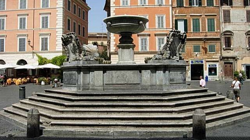 Grande fontana in pietra con vasca centrale su base a gradoni e sculture laterali, collocata in una piazza lastricata. Sullo sfondo si vedono alti edifici storici con facciate color ocra e persiane, oltre a un’area con tavolini all’aperto.