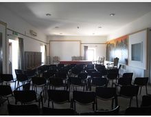 Sala riunioni con file di sedie nere rivolte verso un palco con tavolo e grande schermo di proiezione. Pareti chiare con finestre laterali e tende, armadio in legno a sinistra e grande dipinto murale sulla parete destra.