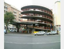 Parcheggio multipiano a rampa elicoidale con grandi solai circolari in cemento e ringhiere arancioni, affacciato su una strada urbana. Alla base si vede un ingresso coperto con pilastri inclinati, con edifici residenziali e auto parcheggiate intorno.