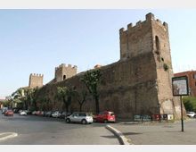 Lungo tratto di mura medievali in mattoni con torri merlate, affacciato su una strada cittadina con auto parcheggiate e alberi allineati. Sullo sfondo si vedono edifici urbani e un cartello stradale vicino al marciapiede.