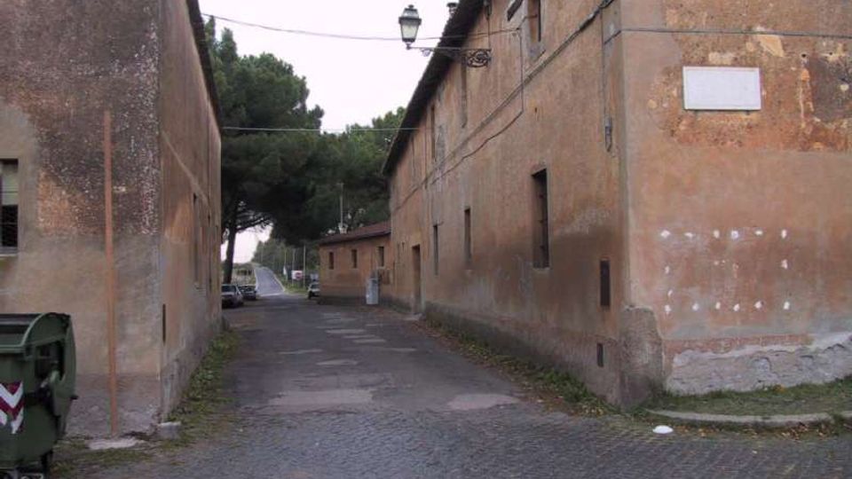 Strada stretta tra due lunghi edifici in intonaco marrone dall’aspetto rurale, con piccole finestre e cavi aerei, illuminata da un lampione sospeso. La carreggiata è in parte acciottolata e conduce verso alberi alti e una strada principale sullo sfondo.
