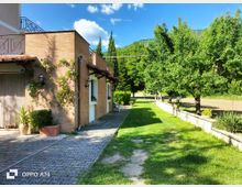 Casa in mattoni con portico e finestre affacciate su un vialetto pavimentato e un prato verde. A destra si vede un frutteto con alberi e un basso muretto, con colline boscose sullo sfondo sotto un cielo azzurro.