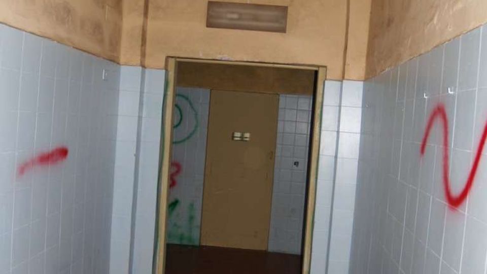 Corridoio interno stretto con pavimento in piastrelle rosse e pareti rivestite da piastrelle azzurre nella parte inferiore e intonaco beige sopra. In fondo c’è un’apertura con porta e un soffitto alto con arco e lampada sospesa, con alcune scritte sui muri.