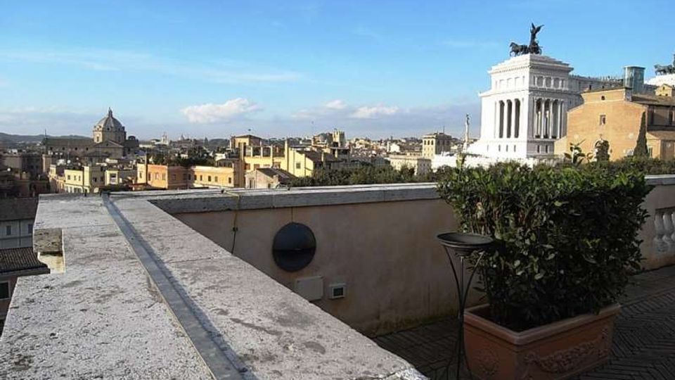 Terrazza panoramica con muretto in pietra e fioriere, affacciata su un ampio skyline urbano. Sullo sfondo si distinguono cupole e un grande edificio monumentale bianco con colonnato e statua in sommità.
