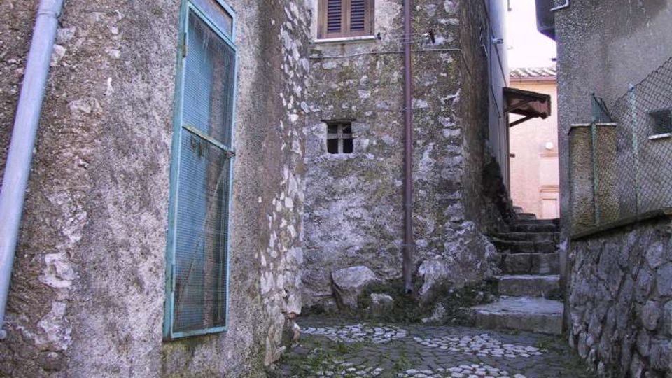Vicolo stretto in un borgo, con pavimentazione in pietra e scalini che salgono tra edifici in muratura grezza. Sulle pareti si vedono una grande porta con grata metallica azzurra, una finestra con scuri e tubazioni a vista; a destra c’è un muretto in pietra con rete metallica.