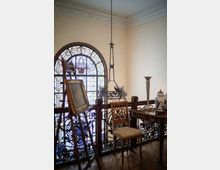 Soppalco interno in stile d’epoca con grande finestra ad arco in vetro colorato e ringhiera in ferro battuto decorata. Nello spazio si vedono un lampadario sospeso, un cavalletto in legno e un piccolo tavolo con sedia e oggetti decorativi.